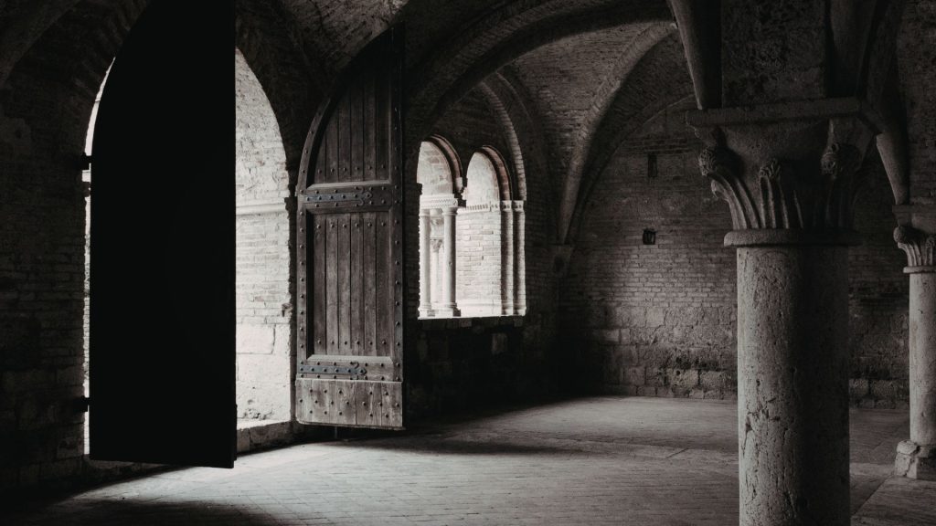 A dimly lit stone hallway with arched ceilings and an open wooden door. Light streams in from outside, casting shadows and creating a moody atmosphere.