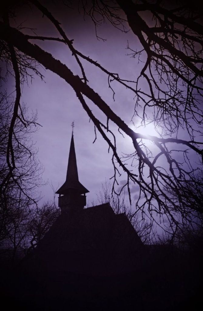 A dark, atmospheric scene features a silhouetted church with a tall steeple against a deep purple sky. Leafless tree branches frame a bright full moon.