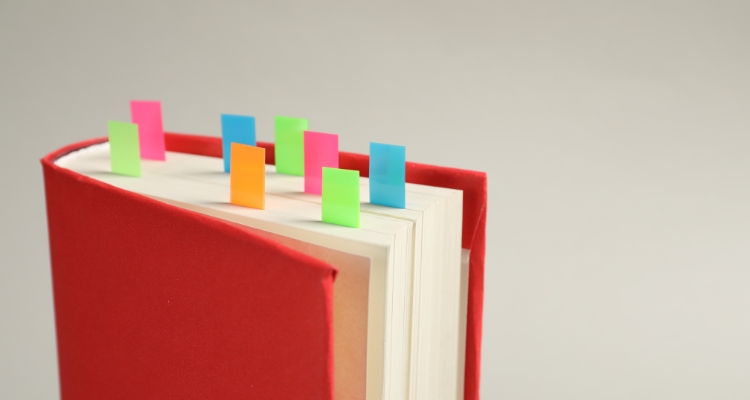 A red hardcover book with colorful sticky tabs protruding from the top, including pink, blue, green, and orange, against a light gray almost white background.