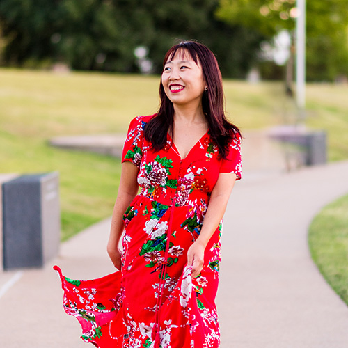 Lyla Lee stands on a paved walkway in a red floral dress, smiling and looking out of frame.