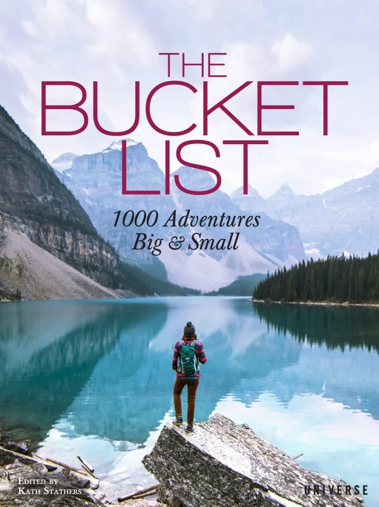 The Bucket List: 1000 Adventures Big & Small by Kath Stathers features a person standing on a rock facing a lake with forest on one side and mountains on the other.