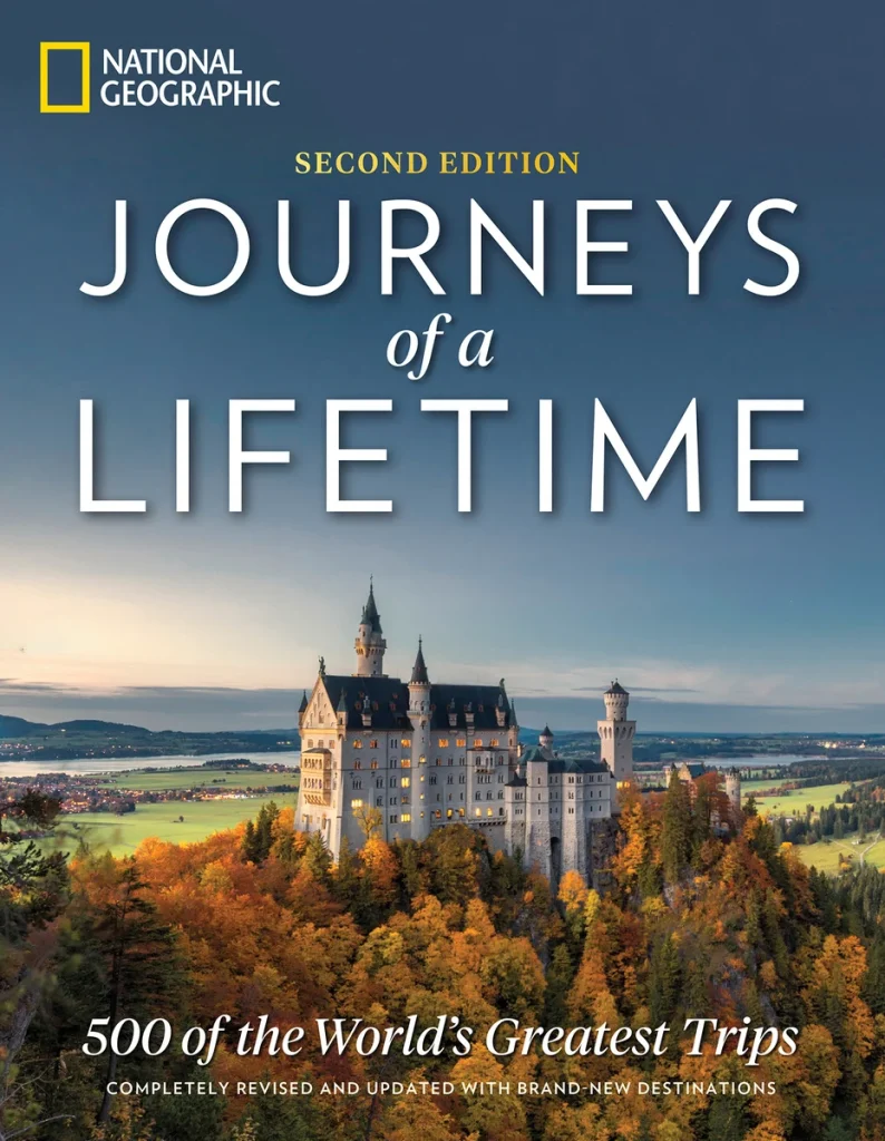 Journeys of a Lifetime: 500 of the World's Greatest Trips by National Geographic features a castle surrounded by a forest with trees of all different colors and a river, and mountains in the background.