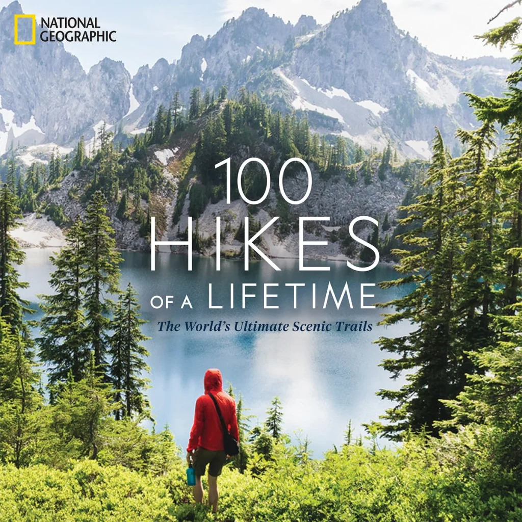 100 Hikes of a Lifetime: The World's Ultimate Scenic Trails by Kate Siber features a person standing on a hiking trail surrounded by trees looking out at the water to more trees and mountains in the distance.