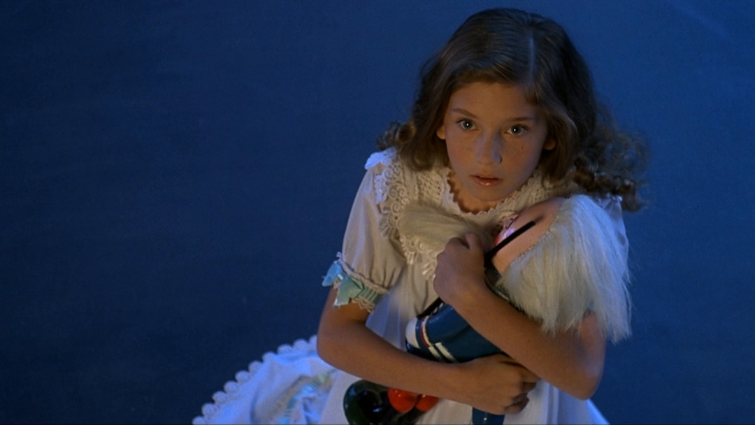 Still from The Nutcracker depicting a young girl with long brown hair, in a white dress, holding onto a nutcracker doll with white hair and a blue coat, looking upwards.