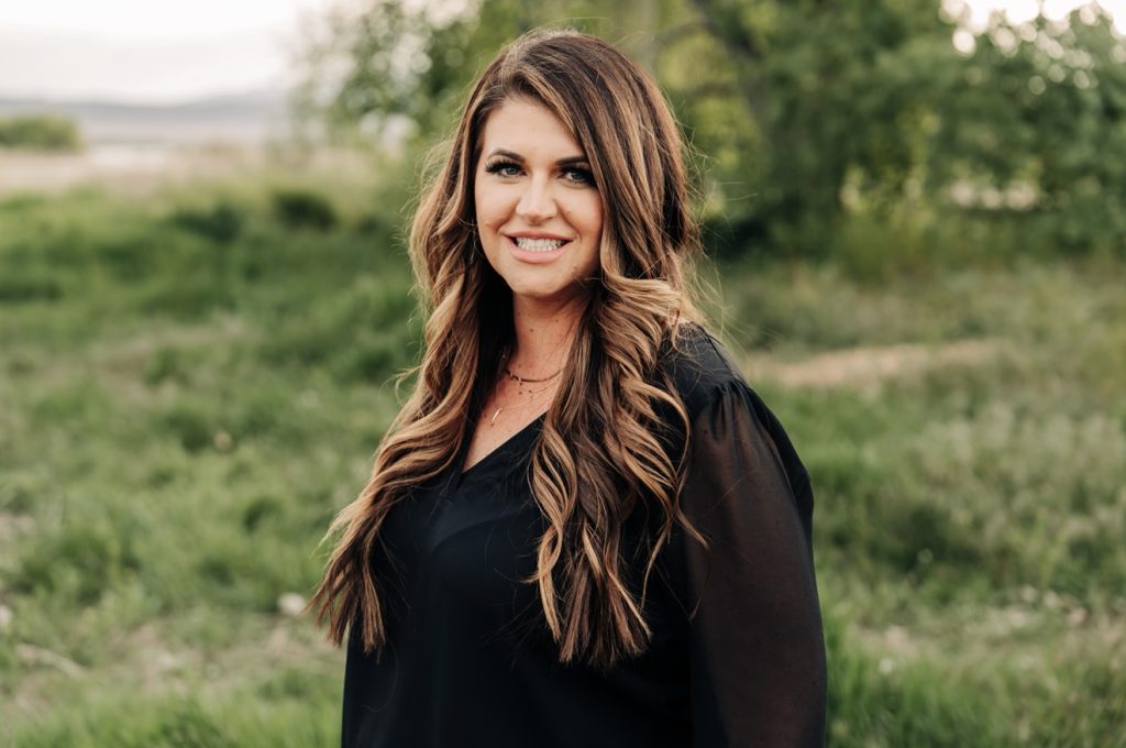 Romance author C.R. Jane smiling at the camera while standing outside against a backdrop of trees and field.