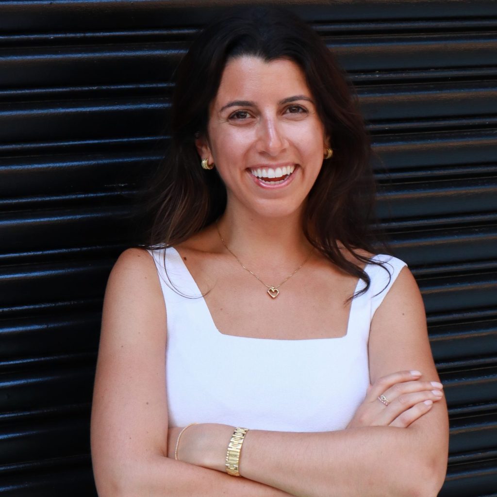 Candice Jalili, a light-skinned Iranian American woman with long dark hair, smiles at the viewer. she wears a sleeveless white top and has her arms folded across her chest. She stands in front of a corrugated metal wall.