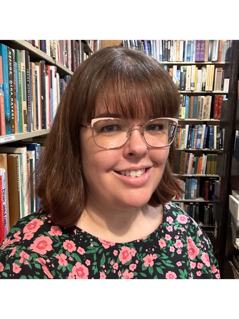 Tabetha of @tabsbooknook, a White woman with short brown hair, horn-rimmed glasses, and a floral top, smiles at the viewer as she stands among a series of bookshelves.