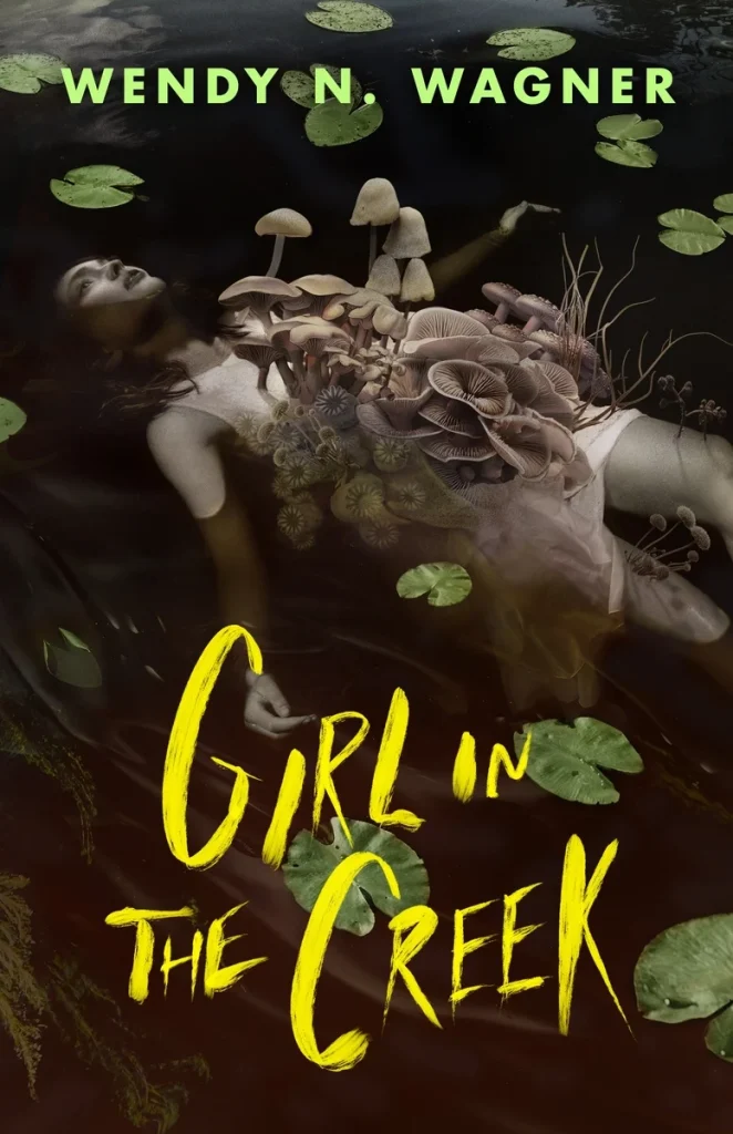 Girl in the Creek, a horror/thriller novel by Wendy N. Wagner, an eerie surreal cover featuring a woman in a white dress lying partially submerged in dark water with mushrooms growing from her chest. Green lily pads float around her as the yellow title text stands out against the shadowy scene.