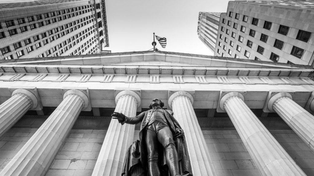 Low angle view of a statue in 18th-century dress standing in front of a building with Greco-Roman columns supporting a triangular roof adorned with an American flag, flanked by towering skyscrapers. The image is black and white.