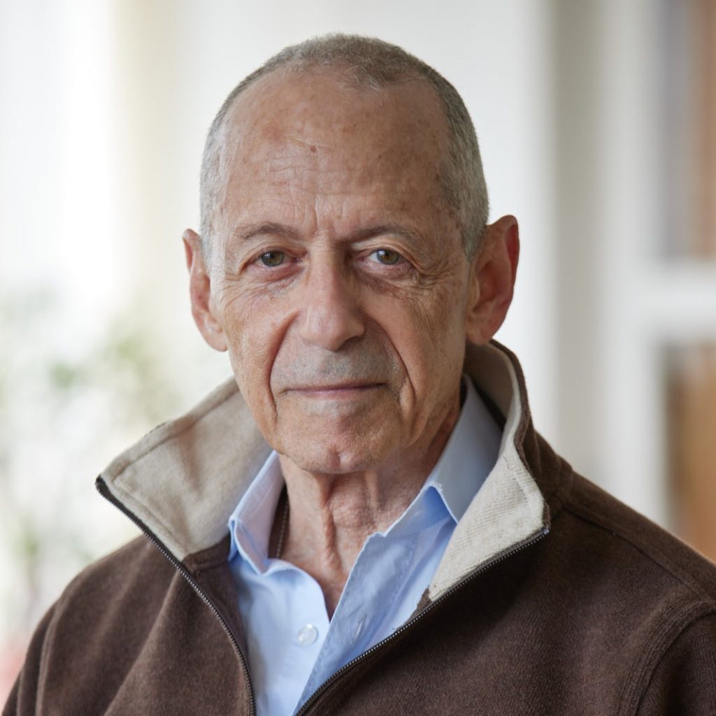 Ken Miller, a bald, older White man in a brown jacket and blue dress shirt, looks at the viewer; a window, bookcase, potted plant, and white wall can be seen out of focus behind him.
