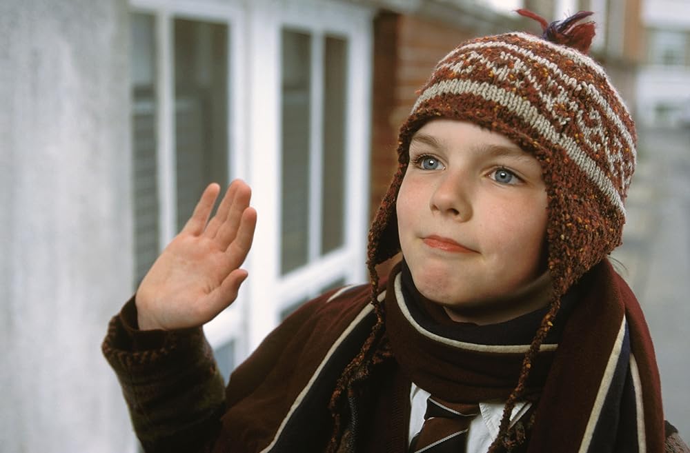 A still from the movie About a Boy. A young boy with pale skin and blue eyes wears an orange knit hat with ear flaps and a white pattern, a maroon scarf, and a brown coat. He stands on a street and waves his right hand in a hello gesture.