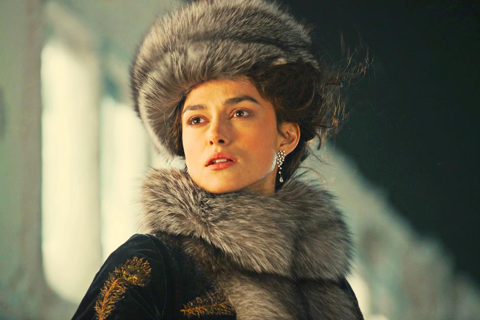 A still from the 2012 movie Anna Karenina. A young woman with pale skin and dark curly hair wears a gray fur hat and collar and a black coat with golden embroidery on the sleeve and expensive diamond earrings. She looks off in the distance with her hair blowing in the breeze.