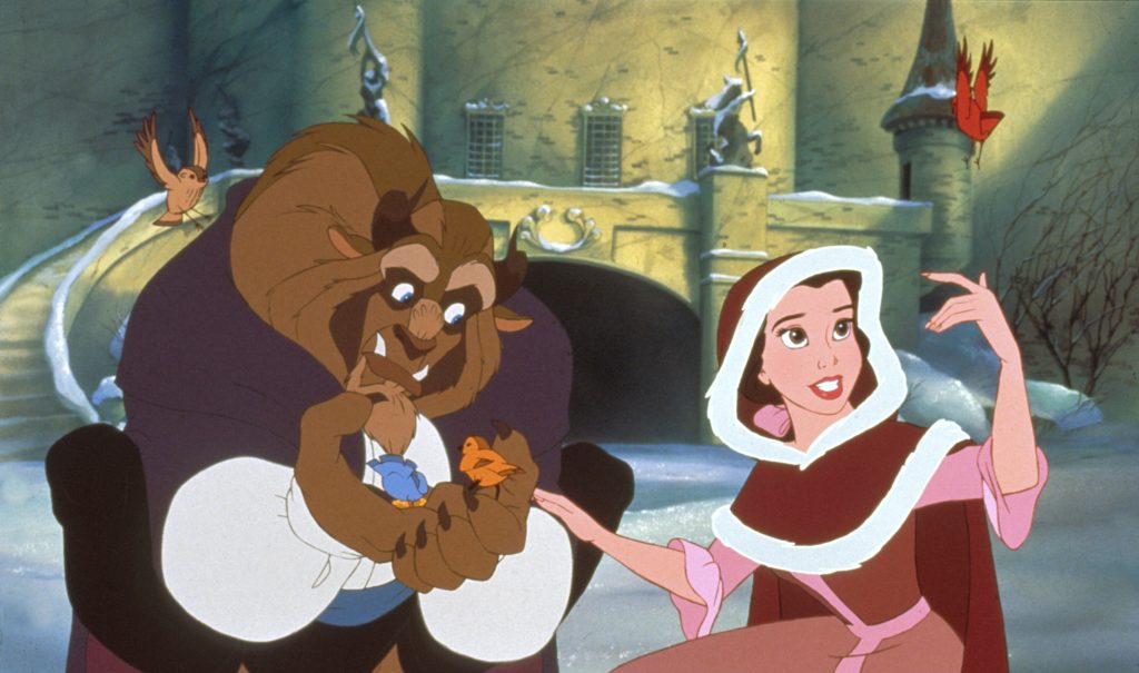 A still from the 1991 animated film Beauty and the Beast. A large beast figure with brown fur and horns wears a white blouse, black trousers, and purple cape. He crouches in the snow and cradles one blue and one orange bird in his hand. A light brown bird is poised to land on his shoulder. To his right, a young woman with pale skin and brown hair wears a pink gown and red cloak lined in white fur. She rests her hand on the Beast's arm and reaches towards a red bird poised to land on her finger. Behind them is a towering castle bathed in golden sunlight.