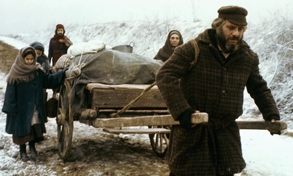 A still from the movie Fiddler on the Roof. An older man with gray hair and a gray and black beard wears a brown hat and brown patterned coat and pulls a handcart full of supplies through a wintry field. Behind him is a woman, two young girls, and a younger man also bundled in cold weather attire.