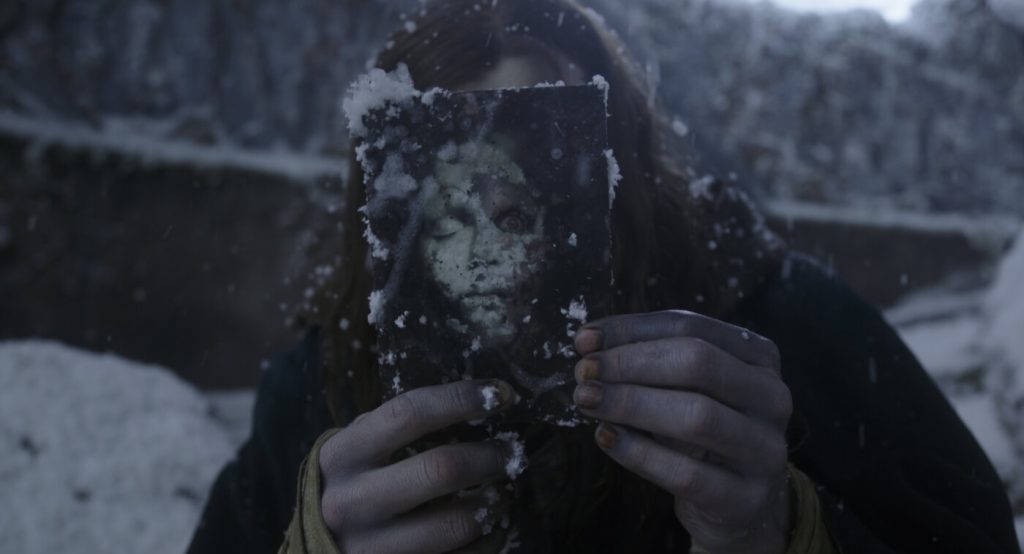 A still from the 2025 movie Frankenstein. A figure with pale skin tinged with blue and long brown hair wears a black cloak and holds an old tintype photograph of a death portrait in front of its face. Behind them is a snowy background.