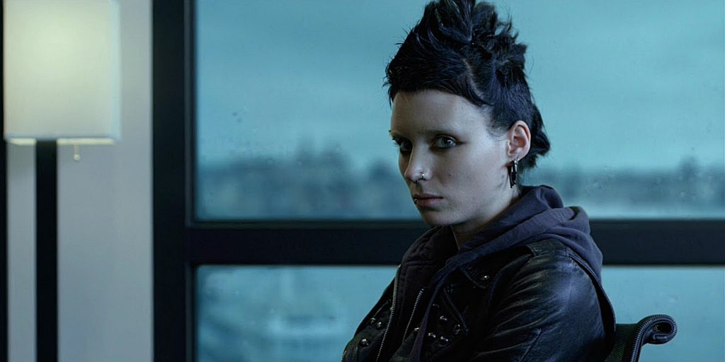 A still from the 2011 movie The Girl with the Dragon Tattoo. A young woman with pale skin, harsh black eyeliner and multiple facial piercings has bleached eyebrows and black hair in a punk rock mohawk. She wears a black leather jacket and sits in a chair in front of a window, looking forward with a guarded expression.
