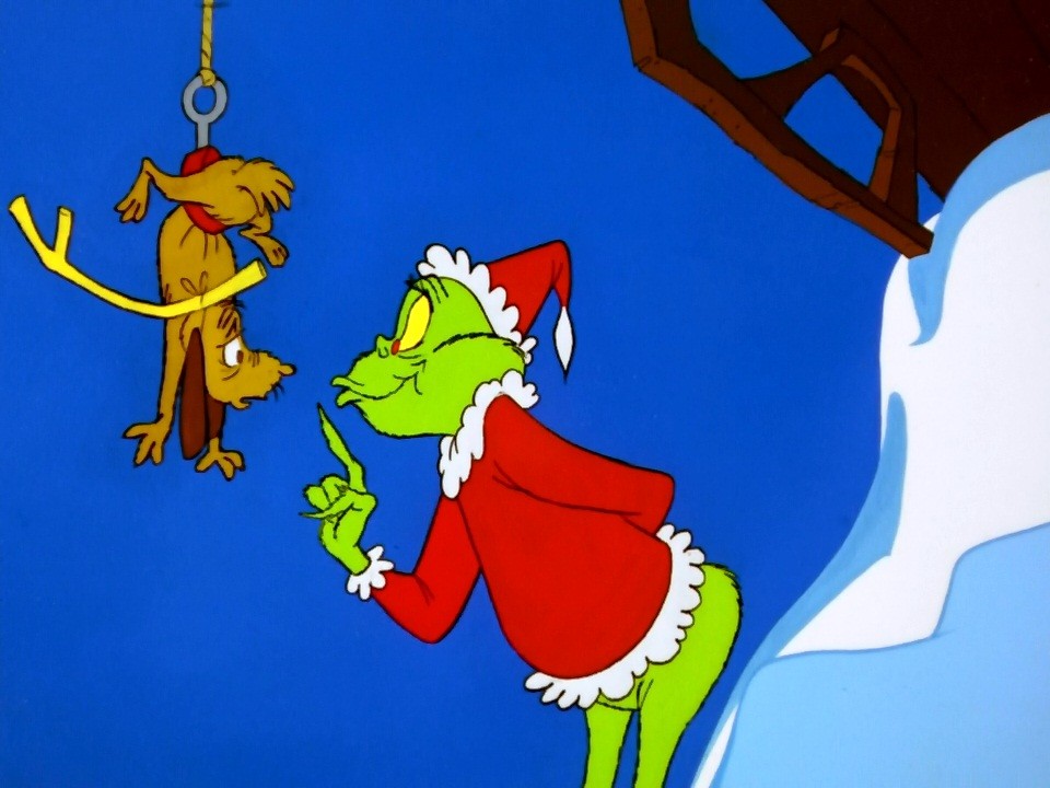 A still from the 1966 movie How the Grinch Stole Christmas! Against a dark blue sky, a creature with fuzzy green fur and yellow eyes wears a red Santa hat and coat trimmed with white fur. He shakes his finger at a small brown dog with an antler tied to its head that is dangling from a dark brown sled resting on a snowy peak.