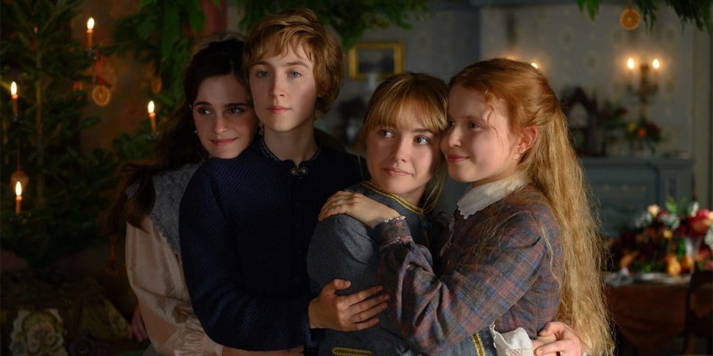 A still from the 2019 movie Little Women. Four young women with pale skin in 19th century dresses hug in a living room decorated with garlands, orange slices, and lighted candles. Behind them is a table with part of a Christmas feast on it.