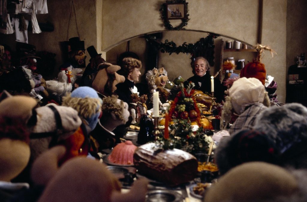 A still from the movie The Muppet Christmas Carol. A table loaded with a Christmas feast is surrounded by muppet characters in Victorian clothing. They all look towards the head of the table where an old man (human) with pale skin and gray hair wears a black suit and smiles. To his left is another human character, a man with pale skin and brown curling hair in a similar black suit.