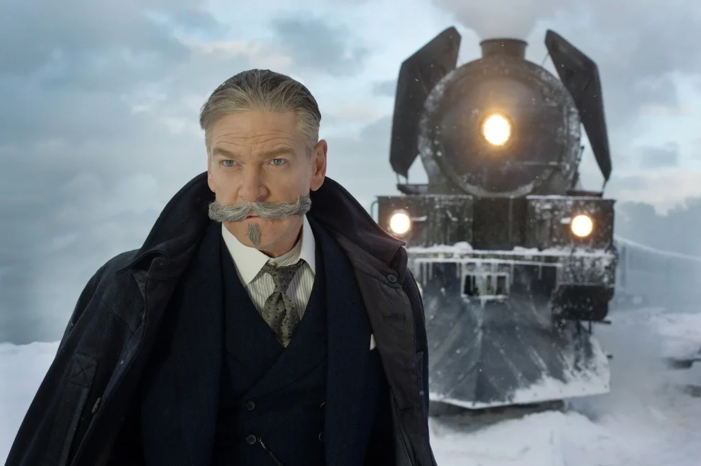 A still from the 2017 movie Murder on the Orient Express. A man wiht pale skin, gray hair, and an elaborate gray mustache and pointed goatee wears a black suit and coat and stares ahead in concentration. Behind him is an iron train in a snowy landscape.
