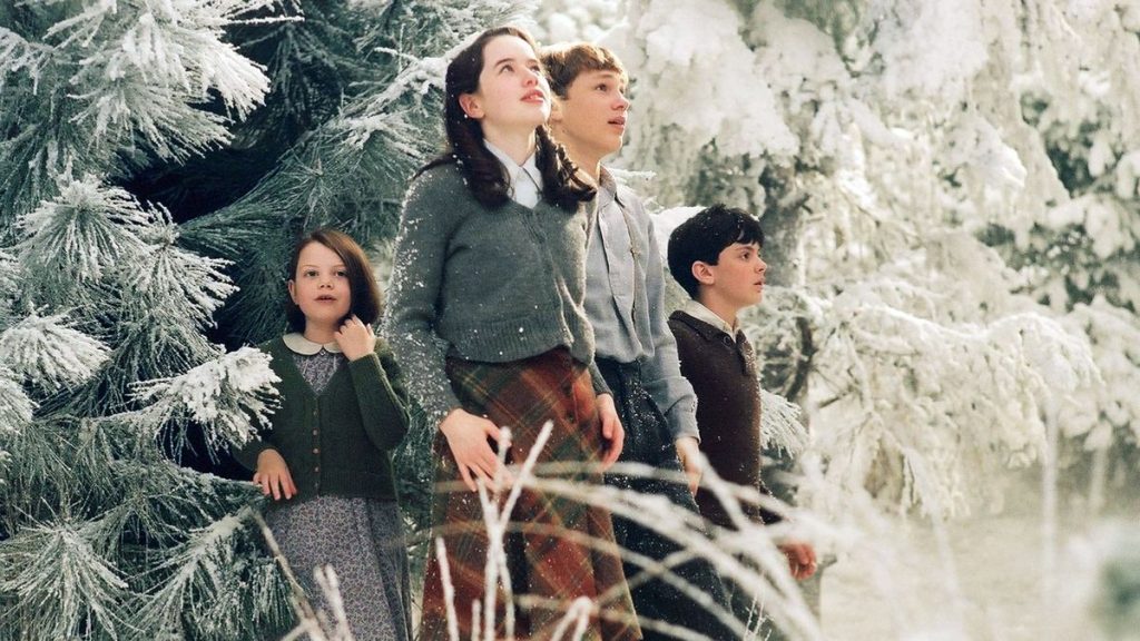 A still from the movie The Chronicles of Narnia: The Lion, the Witch, and the Wardrobe. Four children of various ages wearing old-fashioned clothing stand among snow-covered pine trees and look around in wonder.