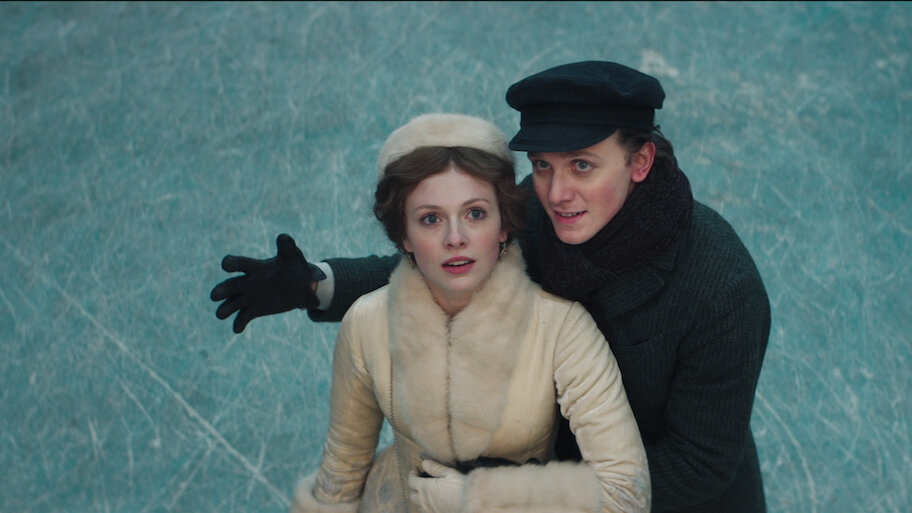 A still from the 2020 movie Silver Skates. On top of a blue, icy surface crisscrossed with skating lines, a young woman with pale skin and golden brown hair wears a white fur coat and hat and stands beside a young man with pale skin wearing a black hat and coat. His arm is wrapped around her and he gestures above them at something they are both looking at off camera.