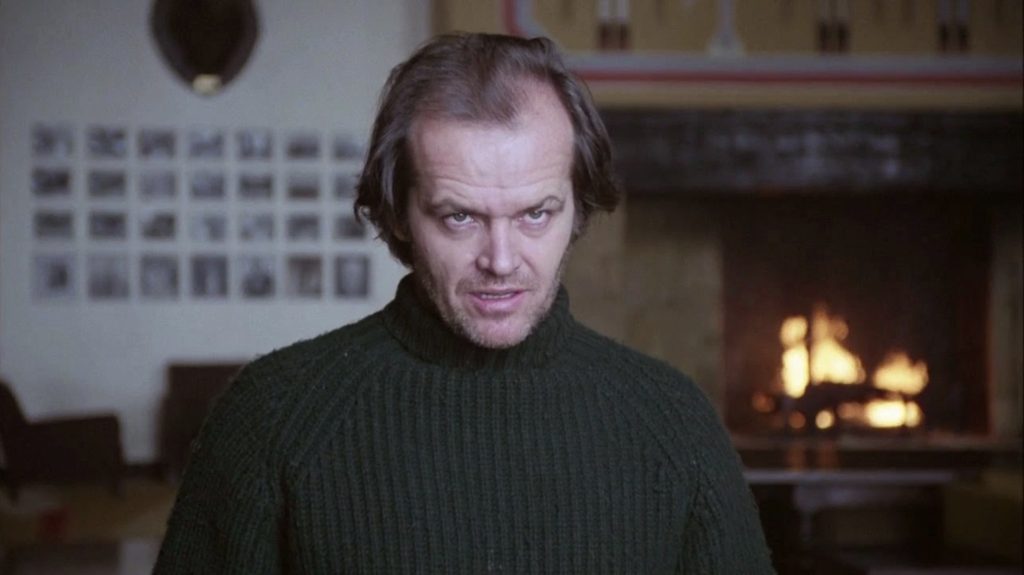 A still from the 1980 movie The Shining. A man with pale skin and brown receding hair and stubble wears a green sweater and stares forward with a menacing expression on his face. Behind him is a large golden brick fireplace and white wall full of old photographs.
