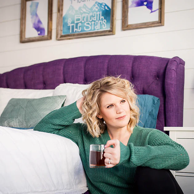 Author Meghan Quinn, a blond haired, light-skinned woman leans against her bed while holding a mug and looking afar.