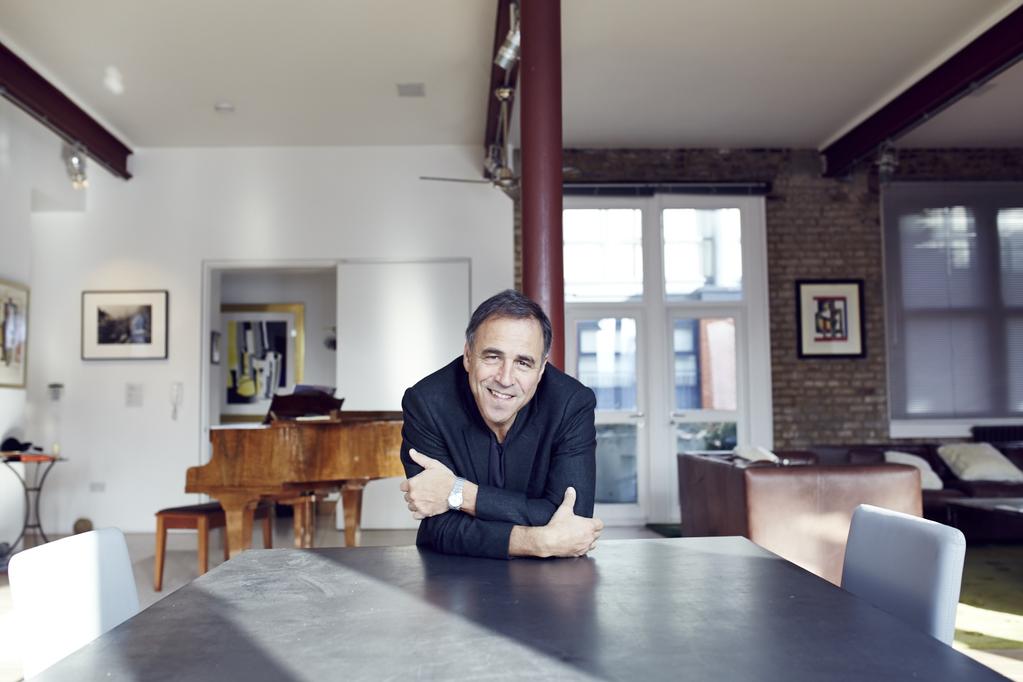 Andrew Horowitz, a man with dark hair wearing a dark blazer, is leaning forward on a black counter with his arms crossed in front of him and smiling at the camera. The background depicts the interior of a house with a brick wall, leather couch, wooden piano, and a white door.