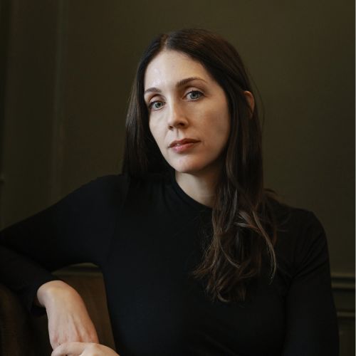 Liz Moore, a woman with long dark brown hair, stares at the camera with her arm resting on a surface beside her. The background is dark brown.