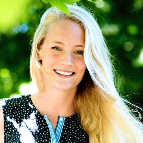 Lucy Foley, a blonde woman with blue eyes wearing a black blouse with white spots, smiles at the camera. The background is blurred but seems to be greenery.
