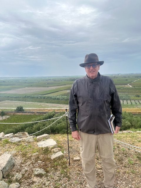 Author James Clark standing beore a field holding two books. He's wearing a fedora and a rain coat in khaki hiking pants.