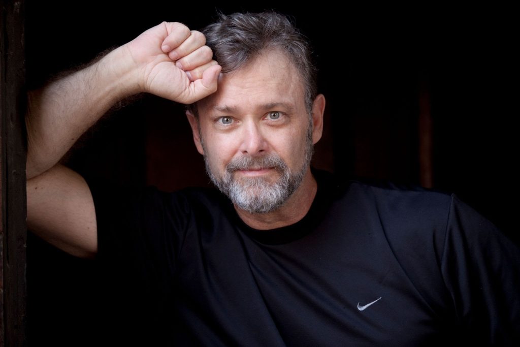 Author Christopher Moore leaning on a wall with his fist at his forehead wearing a black nike t-shirt.