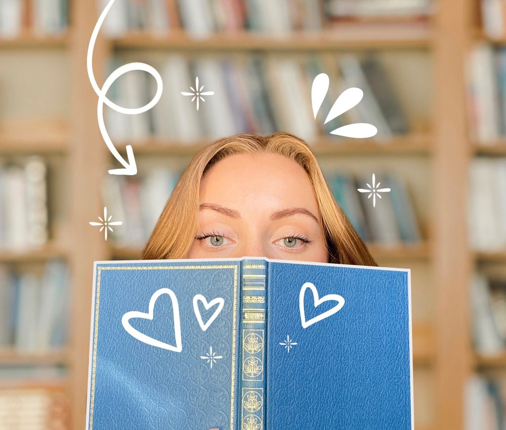 Julie of @flying.reader, a White woman with blonde hair, peers at the viewer from behind an open blue book; an out of focus bookshelf can be seen behind her. The image is decorated with white hearts, sparkles, and a spiraled arrow pointing to Julie's head.