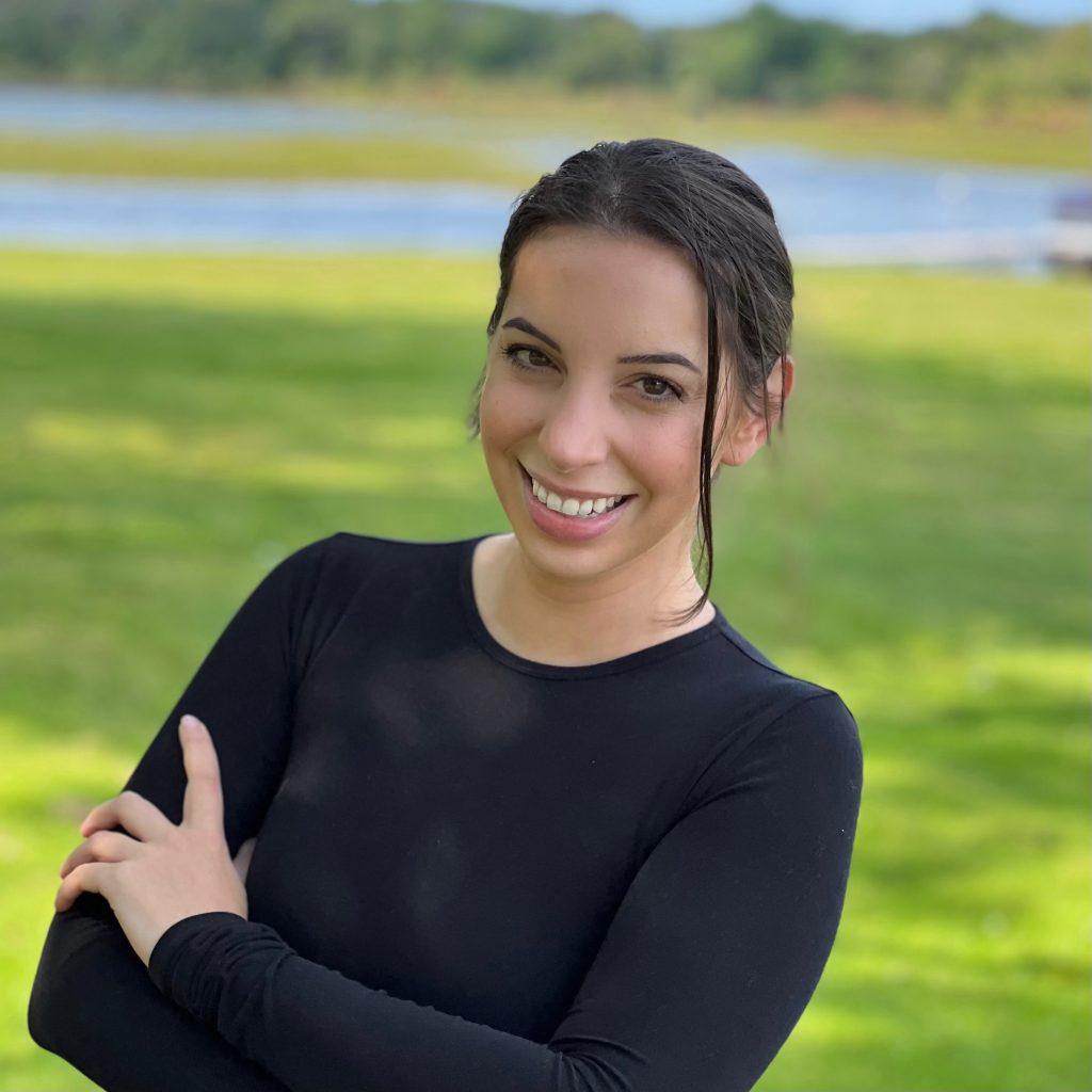Author Ellie K. Wilde stands in front of a pond with green grass as she crosses her arms in front of her. She wears a black crewneck long sleeve shirt and has her brown hair pulled back, a single tendril is out off to the side of her face. She gives the camera a warm smile.