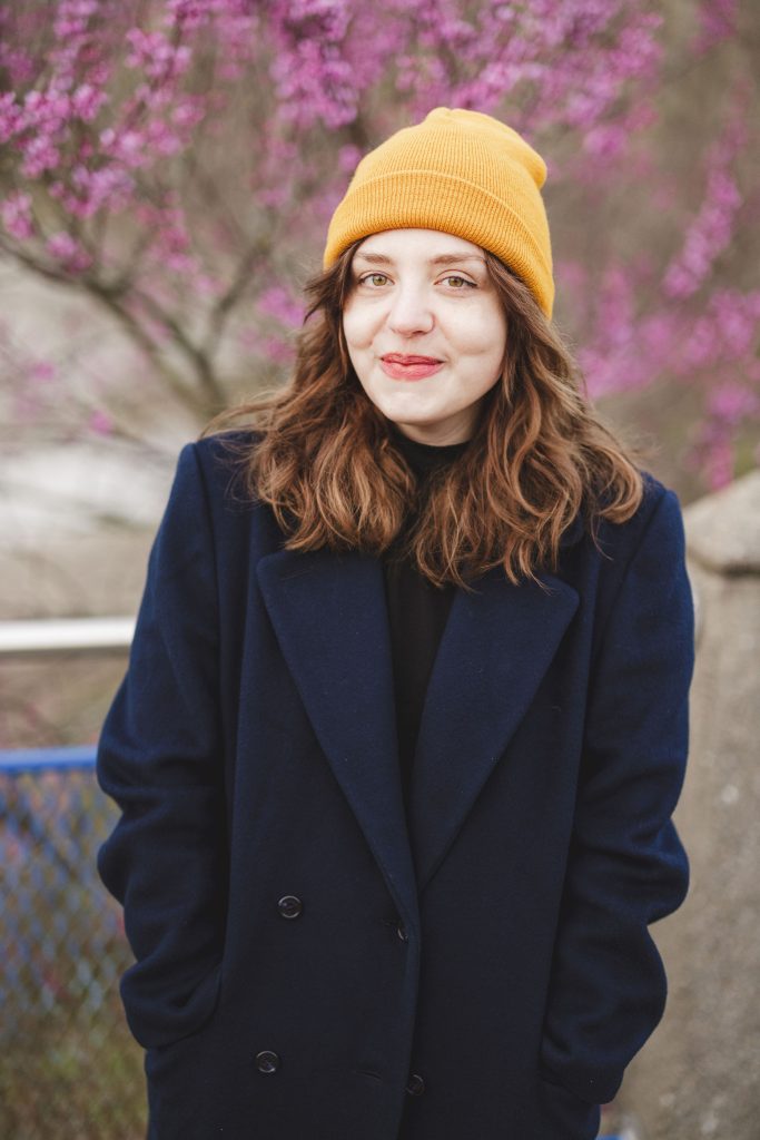 Author Madge Maril's headshot of her looking directly into the camera with a closed-lip smile. Madge wears a vivd yellow beanie with her medium-length brown hair falling around her shoulders in natural waves. She's bundled, hands in pockets, in a dark navy coat with a black shirt underneath.