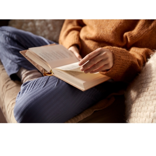 A person in a cozy brown sweater sits cross-legged on a light-colored couch, turning a page in a book.