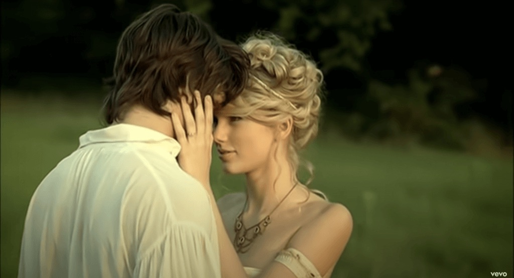 A still from the music video for Love Story by Taylor Swift. Taylor Swift has her blond hair arranged in a curly updo and wears a bronze necklace and a white off-the-shoulder gown. She holds the face of a young man with brown hair wearing a white button down shirt. They stand in a green meadow.