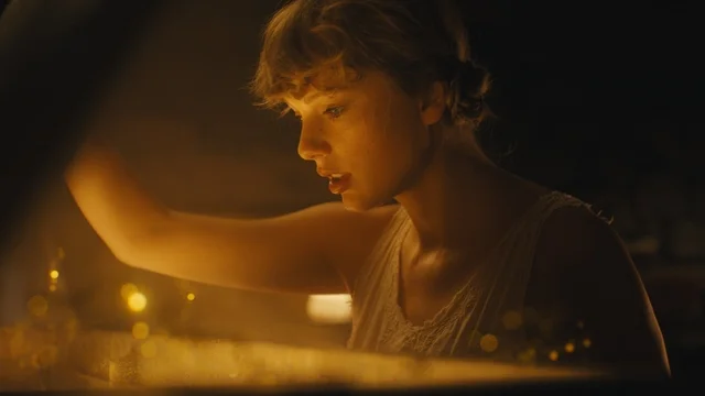 A still from the music video for cardigan by Taylor Swift. Taylor Swift wears a white tank top with crochet detail and looks inside an object filled with golden sparkling light.