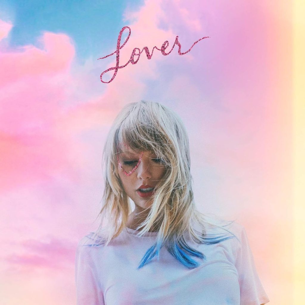 The album cover for Lover by Taylor Swift. Taylor Swift wears a pale tie dyed t-shirt with the ends of her blonde hair dyed blue and a pink glitter heart drawn around her right eye. Behind her is a background of pink, purple, and yellow clouds.