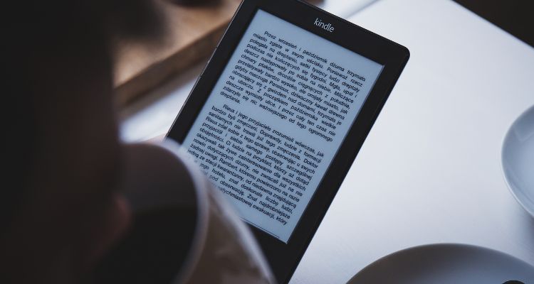 A Kindle eReader being red with a blurry image of someone drinking from a tea cup