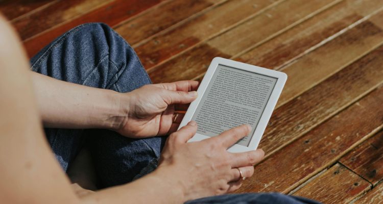 A light skinned person sitting cross legged on a wooden floor holding a white Kindle