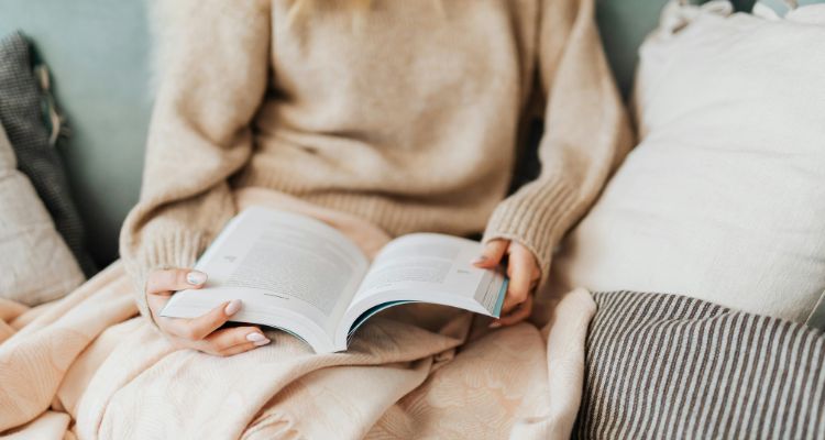 Pictures a girl in a tan sweater reading a book on a couch with a blanket on. Her head is not pictured, it is just her torso.