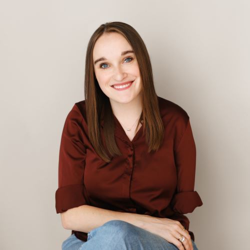 Tennessee Hill, a White woman with long brown hair and a red button-down shirt, smiles at the viewer. She is sitting in front of an off-white background.