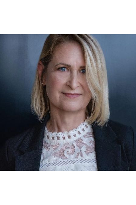 Emily Carpenter, a White woman with chin-length blonde hair, a white lacy top, and a black jacket, looks at the viewer; the background is a gray gradient.