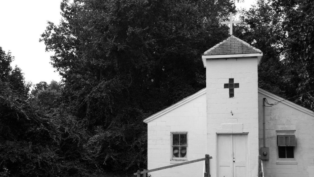 A small white church surrounded by trees with a cross window; the image is black and white.