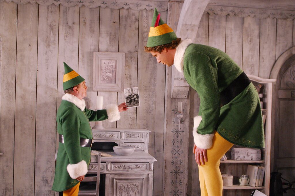 A still from the movie Elf. An elf with pale skin and brown hair wears a green and yellow elf costume trimmed with white fur. He holds out a black and white photograph to a tall human man wearing an identical elf costume hunching over to avoid the low ceiling. They stand in a white-washed wooden room with blue snowflake designs on the beams.