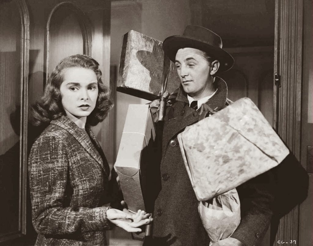 A black and white still from the 1949 movie Holiday Affair. A man wearing a dark coat and hat stands in an open doorway holding a collection of gift wrapped packages and gazing adoringly the woman beside him. The woman has soft wavy hair and a tweed jacket and looks away in concern.