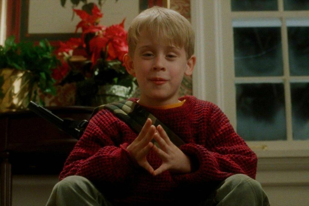 A still from the movie Home Alone. A young blonde boy with pale skin wears a red sweater and sits with a mischievous look on his face. He has a BB gun with a camo strap slung across his back. Behind him is a small table with red poinsettias.