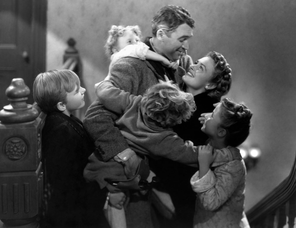 A black-and-white still from the movie It's a Wonderful Life. A smiling man wearing a winter coat is surrounded and hugged by four young children. He gazes down at a woman that looks up at him adoringly.